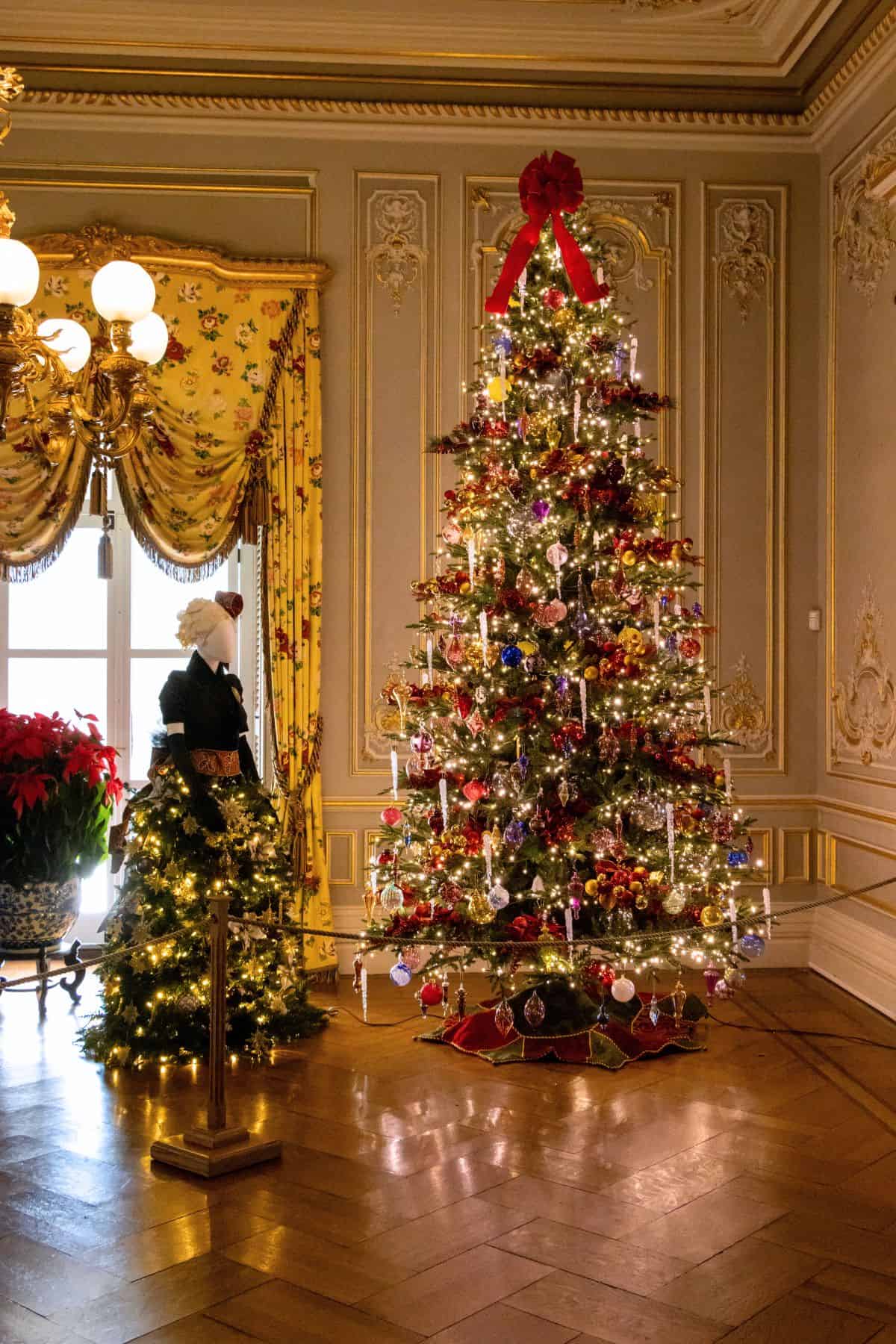 A decorated Christmas tree with lights and ornaments stands in an ornate room with gold accents, next to a smaller tree and a mannequin dressed in historical costume.