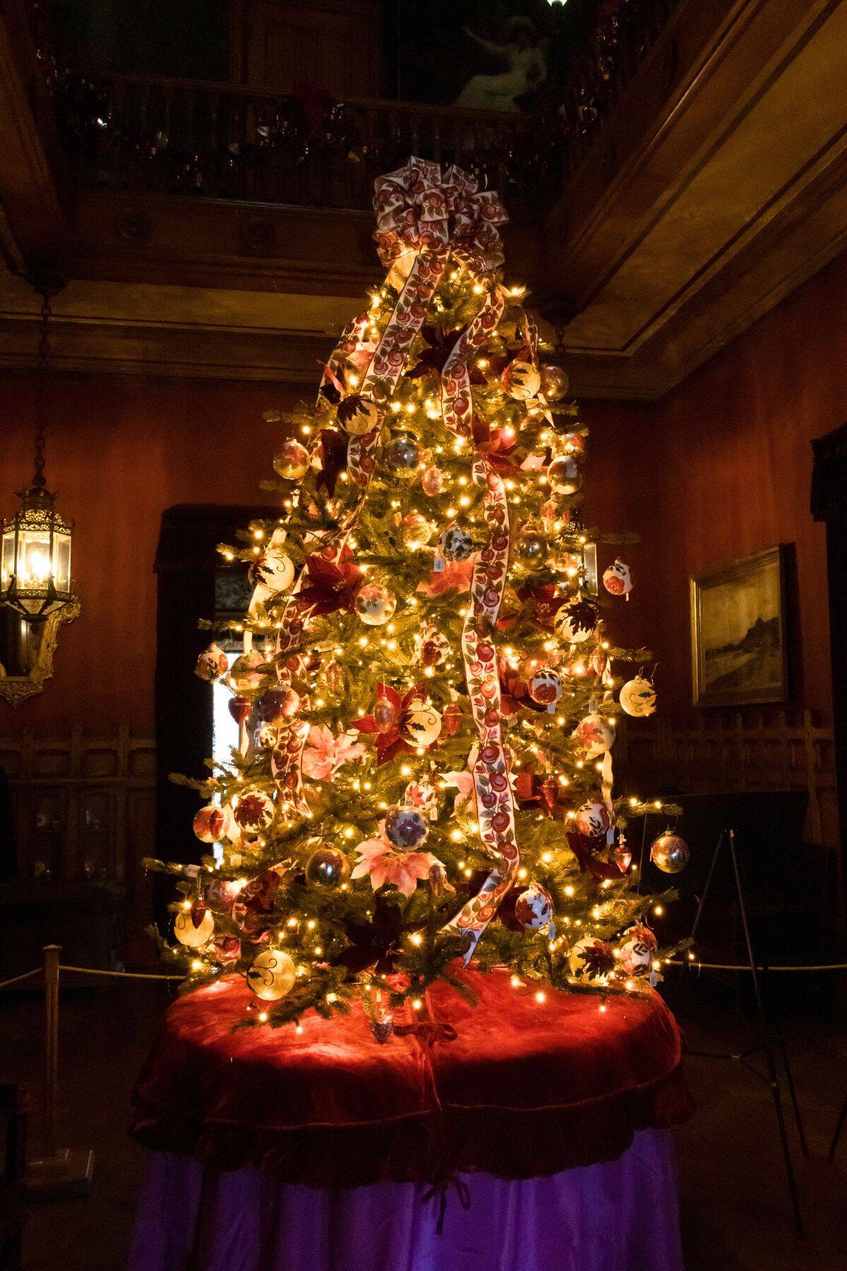 A decorated Christmas tree with lights, ornaments, and ribbon stands on a red and purple-draped platform in a dimly lit, ornate room.
