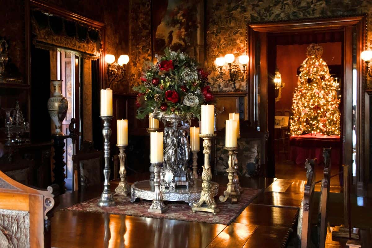 A formal dining room with a long wooden table, ornate candle holders, a floral centerpiece, and a decorated Christmas tree visible in the next room.