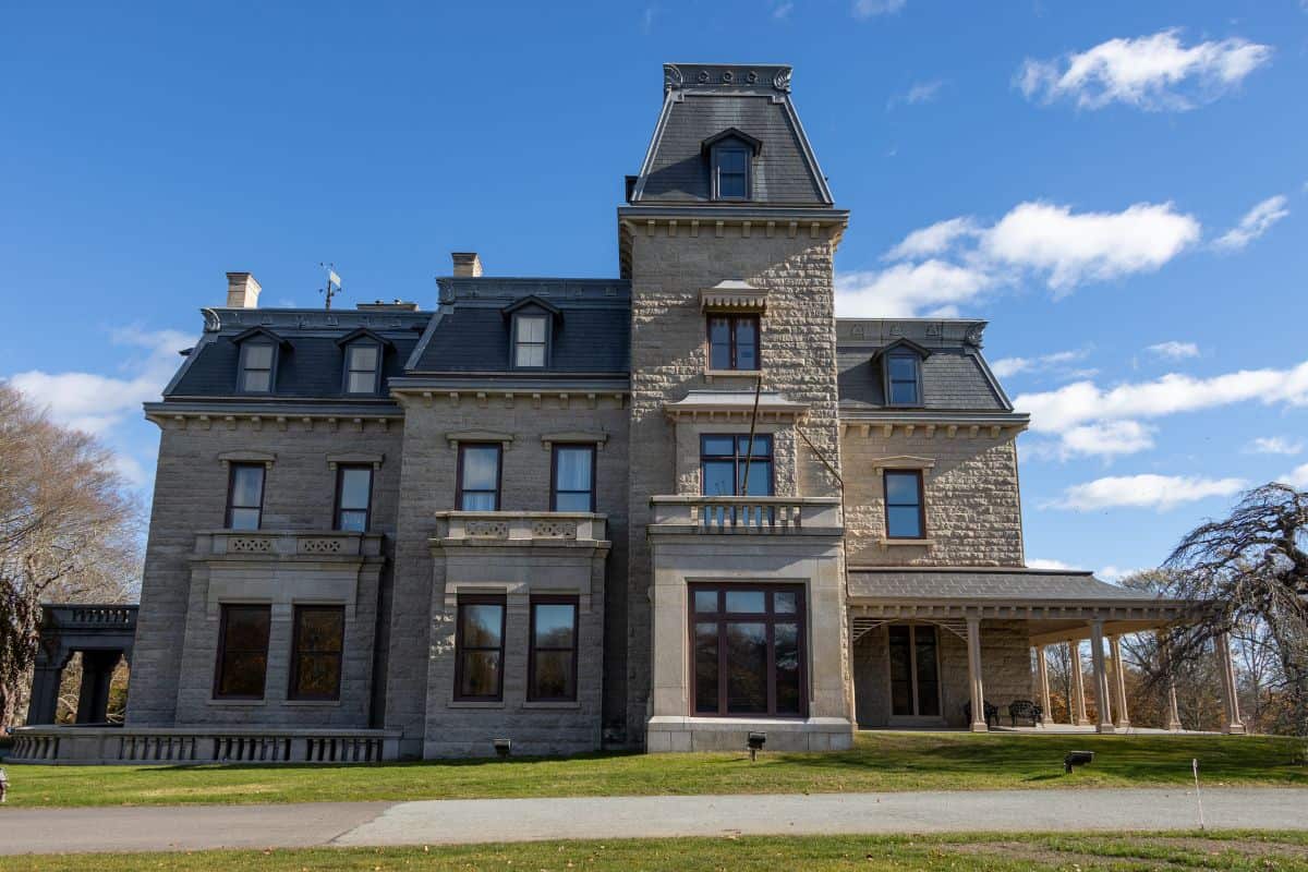 Large stone mansion with a mansard roof, multiple chimneys, and large windows, set on a grassy lot under a blue sky with scattered clouds.