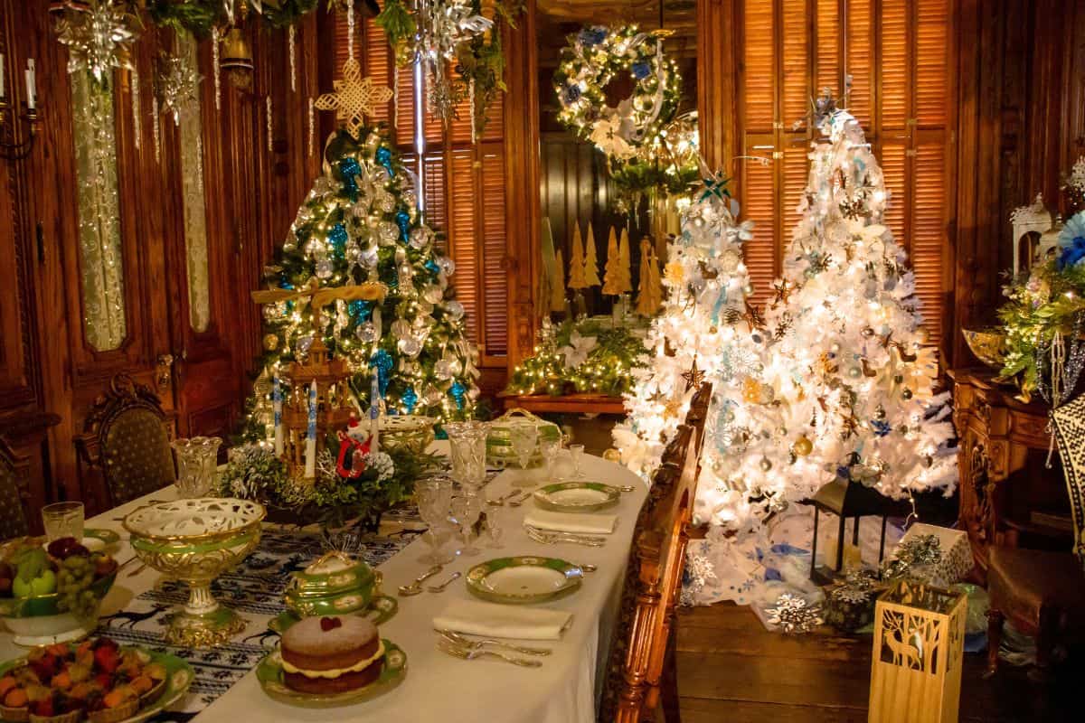 A dining room decorated for Christmas with a set dining table, green and white dishware, food, and three lit Christmas trees adorned with ornaments and lights.