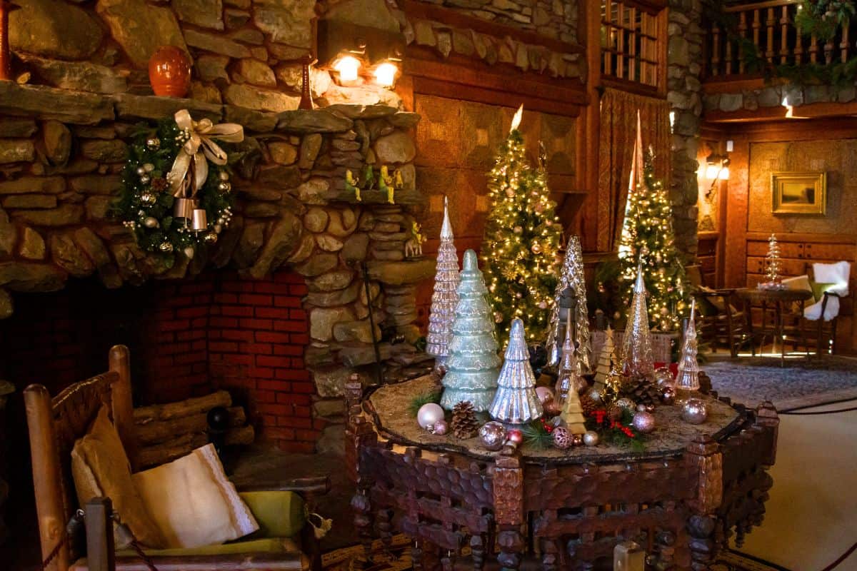 A rustic living room decorated for Christmas with a stone fireplace, wreaths, lit Christmas trees, and an arrangement of glass tree ornaments on a wooden table.