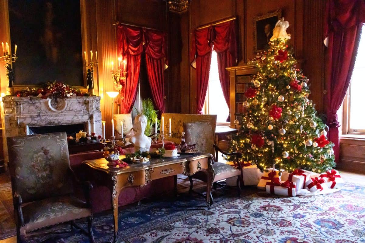Elegant room decorated for Christmas with a lit tree, wrapped gifts, red curtains, a marble fireplace, candles, and antique furniture on a patterned rug.
