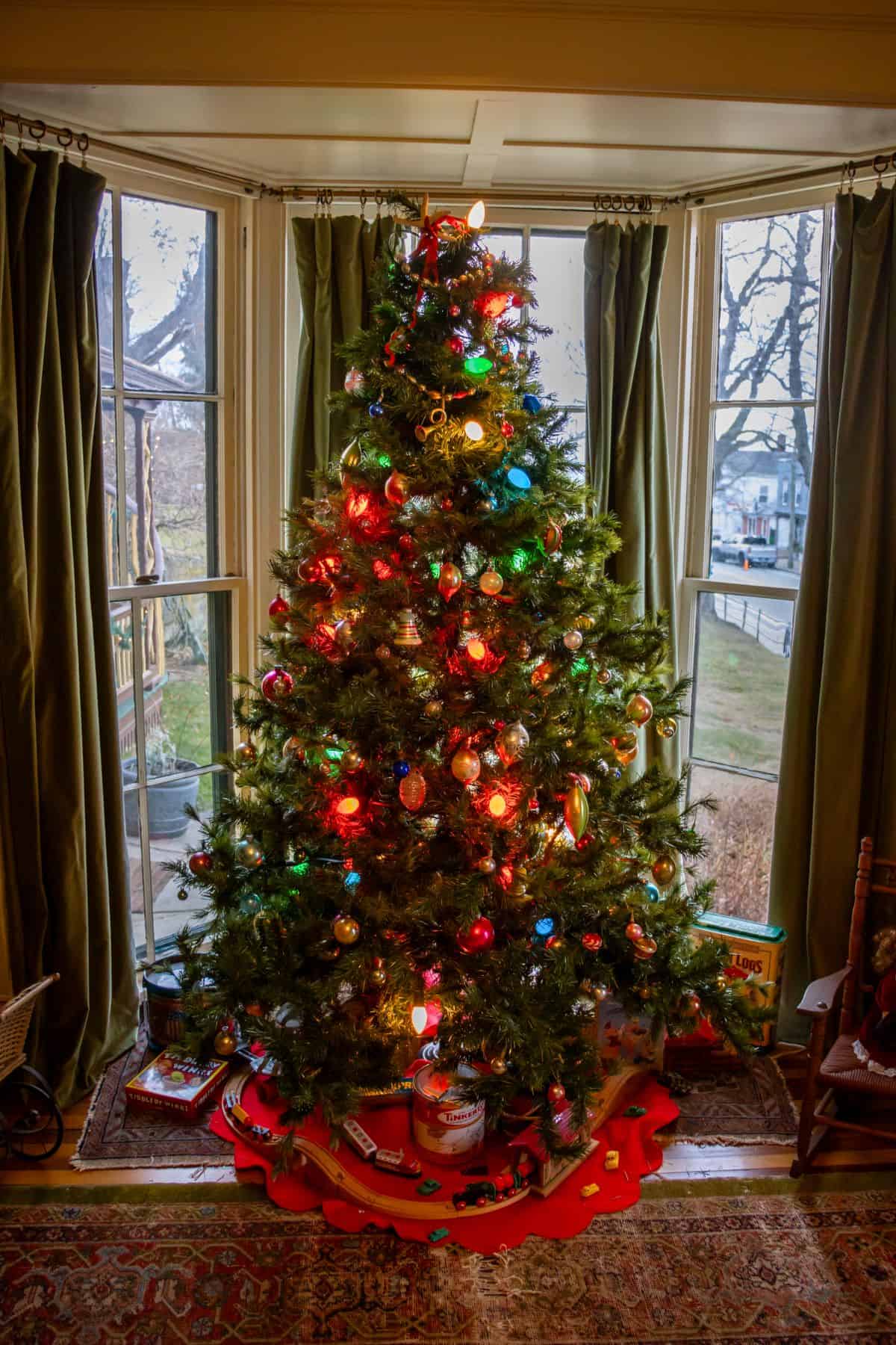 A decorated Christmas tree with multicolored lights and ornaments stands in front of a window with green curtains, with presents placed underneath.
