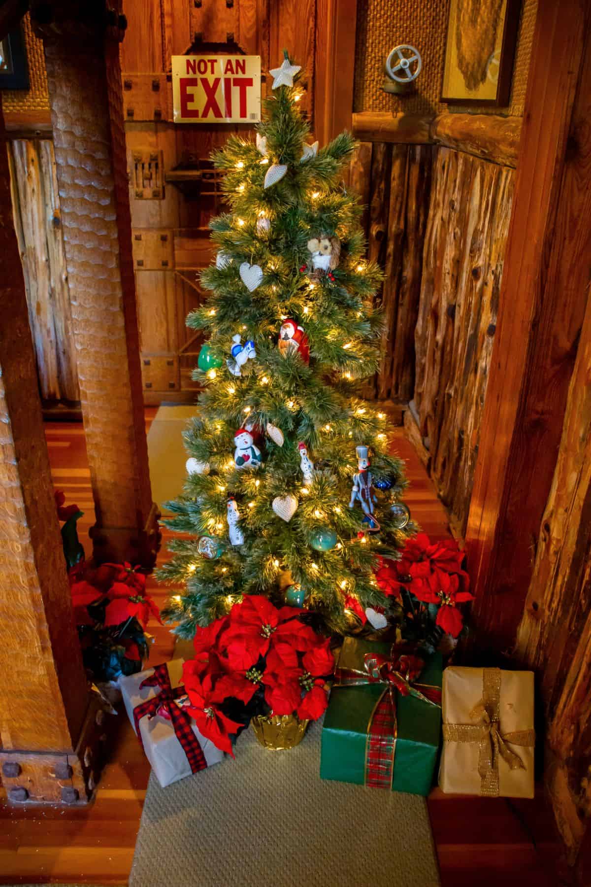 A decorated Christmas tree with lights and ornaments stands indoors, surrounded by wrapped gifts and red poinsettias. A "Not an Exit" sign is visible on the wall behind the tree.