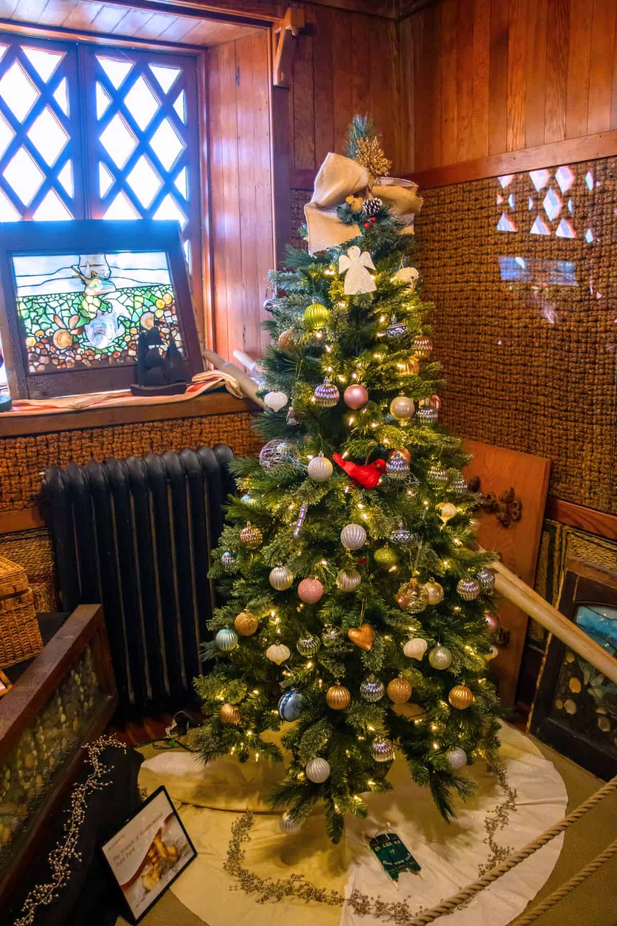 A decorated Christmas tree with lights and ornaments stands in a wood-paneled room near a window, surrounded by various framed artworks and other decor.
