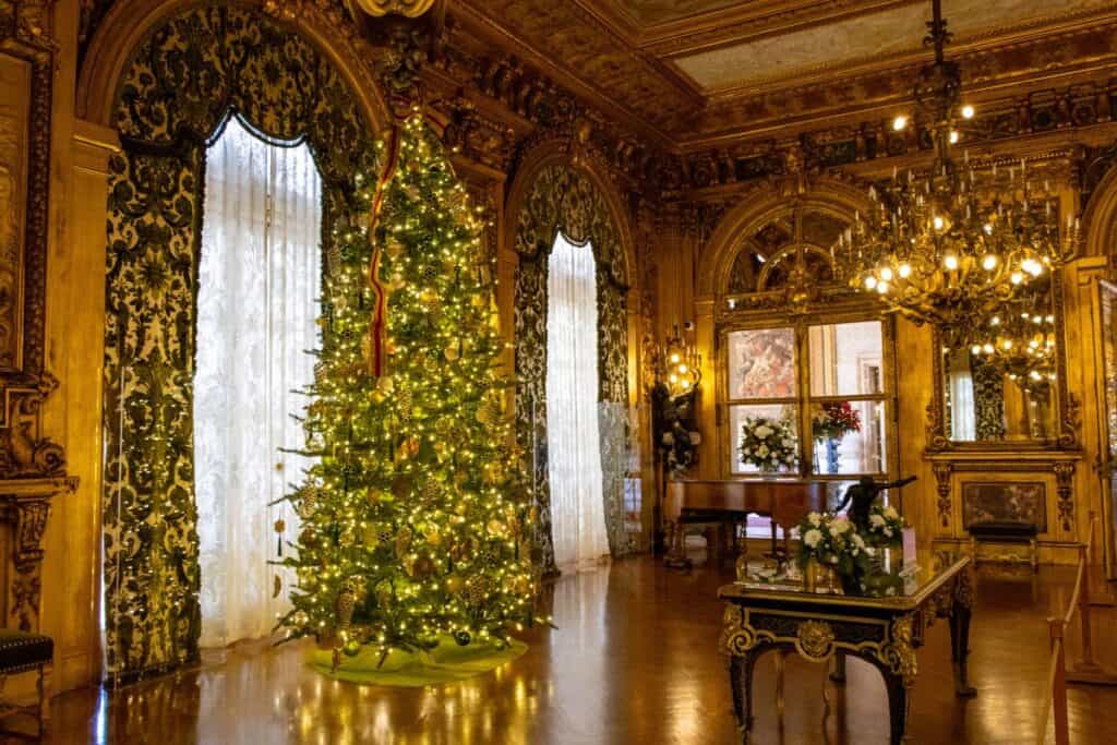 A decorated Christmas tree stands in an ornate, gold-accented room with chandeliers, large windows, and polished wooden floors.