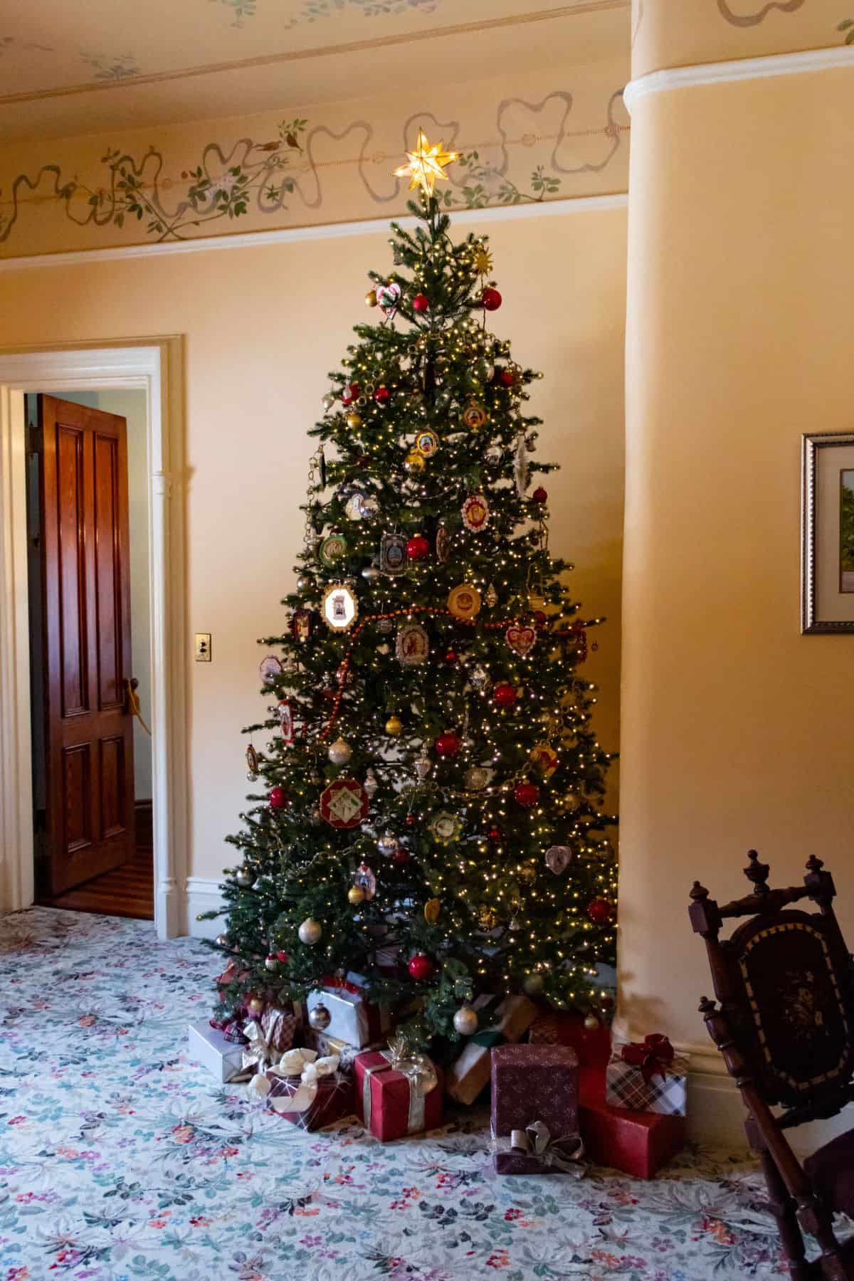 A decorated Christmas tree with lights and ornaments stands in a living room, surrounded by wrapped gifts on a floral carpet.
