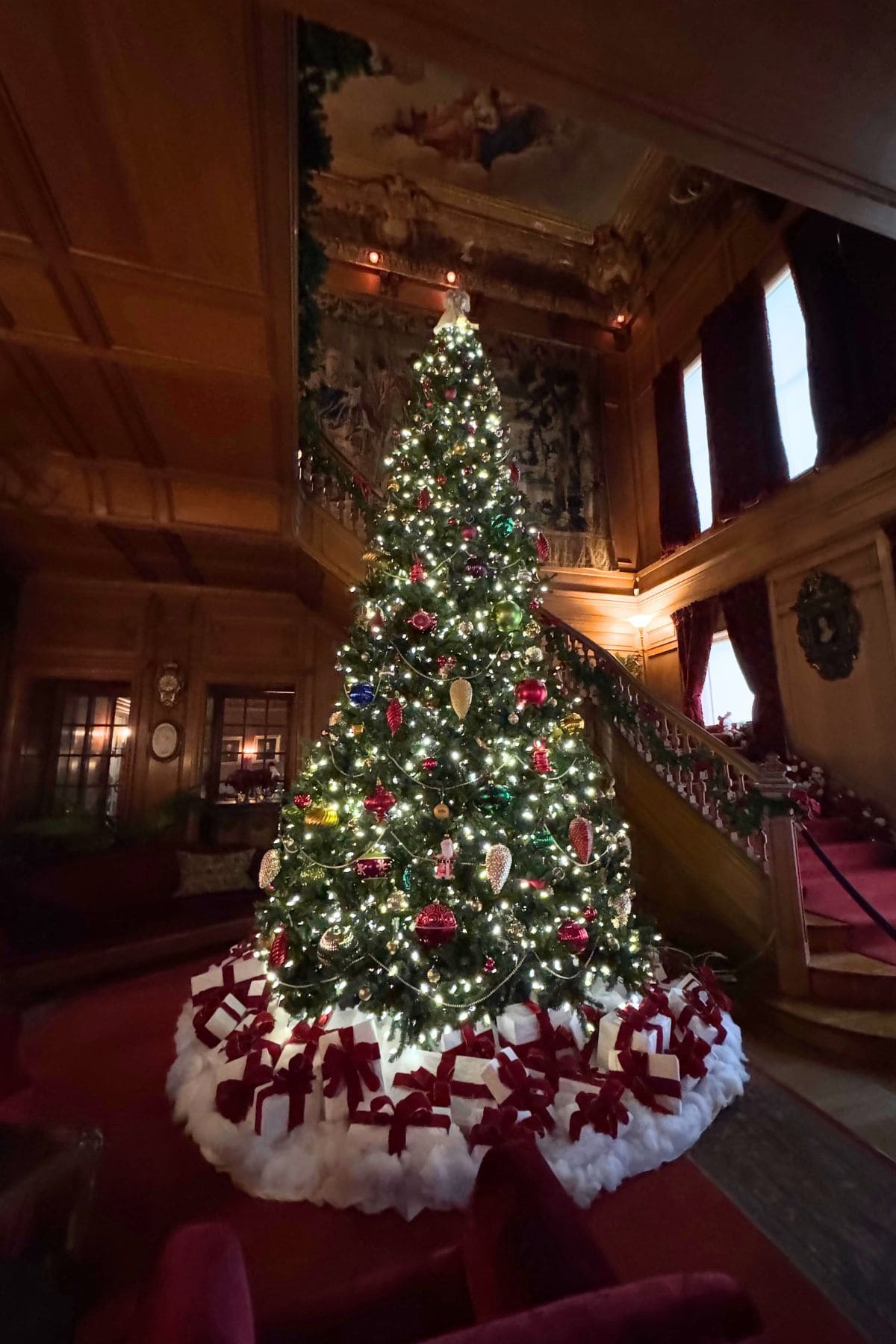 A decorated Christmas tree with lights and ornaments stands in a grand room with wood paneling, red carpet, and a staircase in the background.