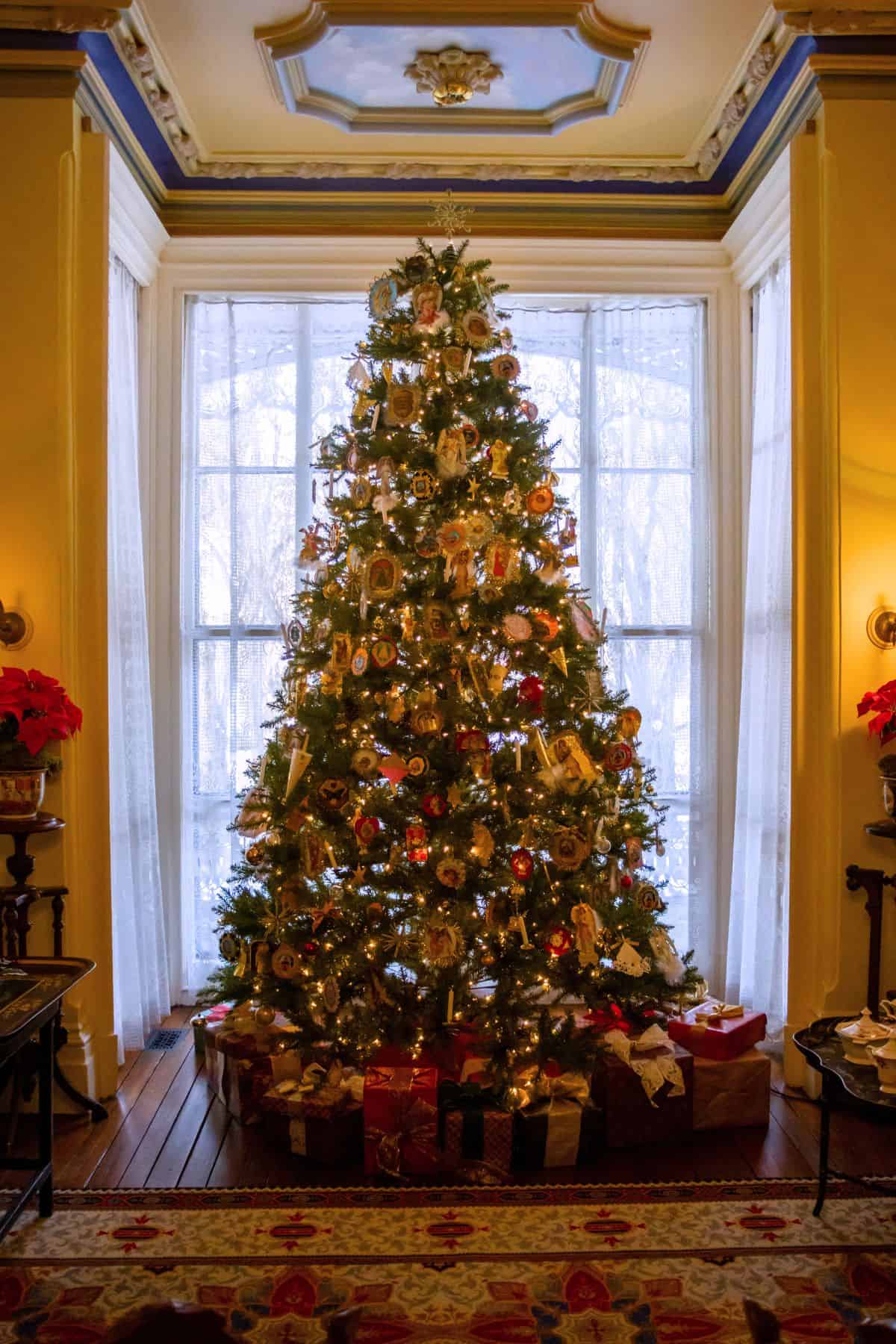 A decorated Christmas tree with lights and ornaments stands in front of a large window, surrounded by wrapped presents on a wooden floor in a warmly lit room.
