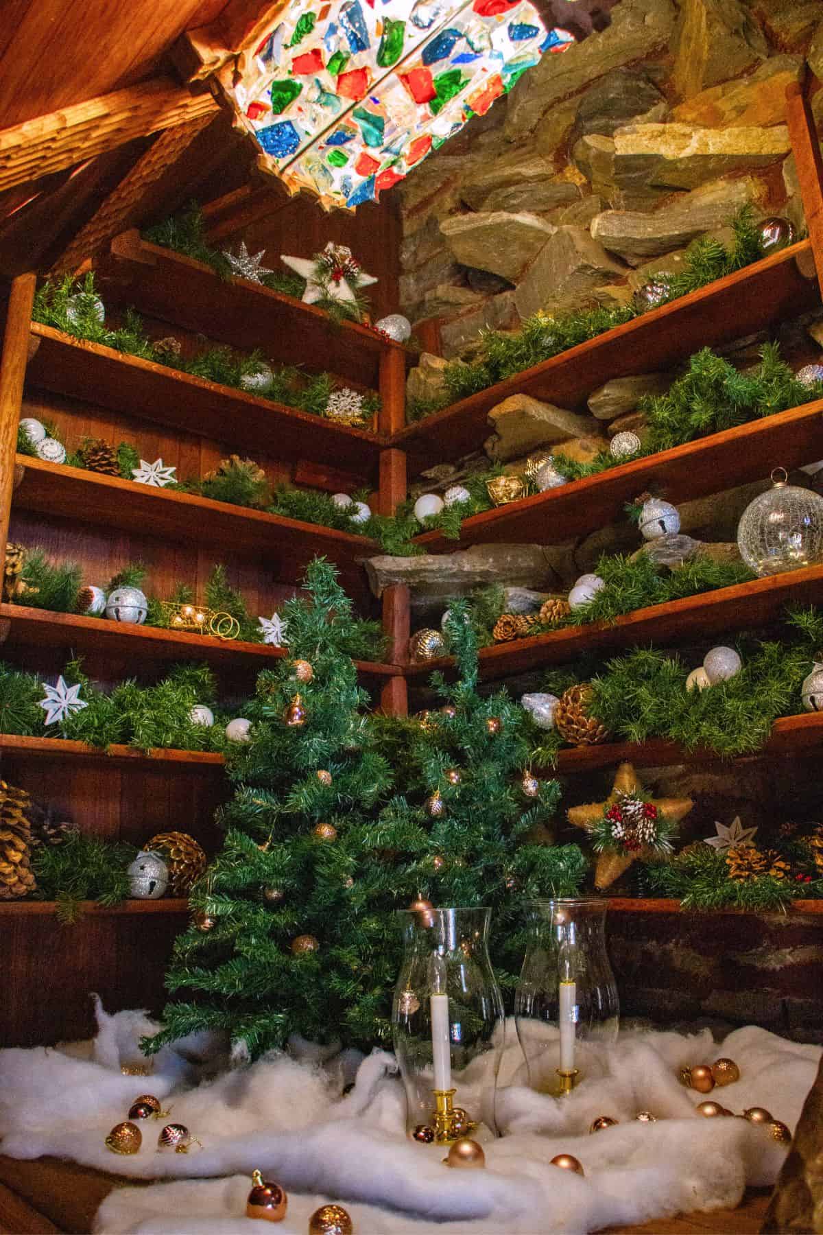 Corner shelves decorated with greenery, ornaments, and pinecones; two small artificial Christmas trees, faux snow, candles, and gold baubles on the floor; stone wall in background.