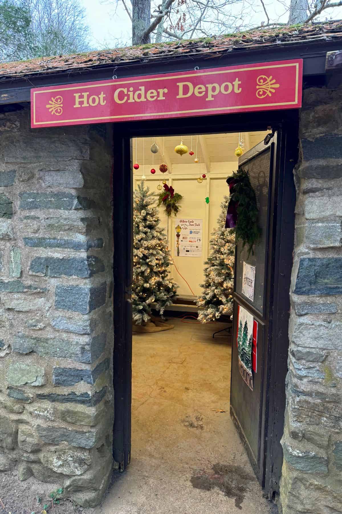 Stone building with an open door under a sign reading "Hot Cider Depot." Inside, decorated Christmas trees and holiday ornaments are visible.