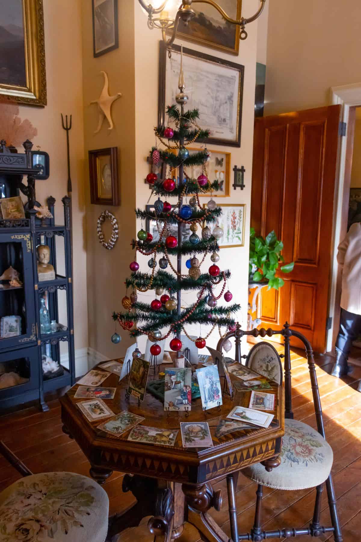 A small decorated Christmas tree with ornaments and cards sits on a round wooden table in a warmly lit room with antique furniture and artwork.