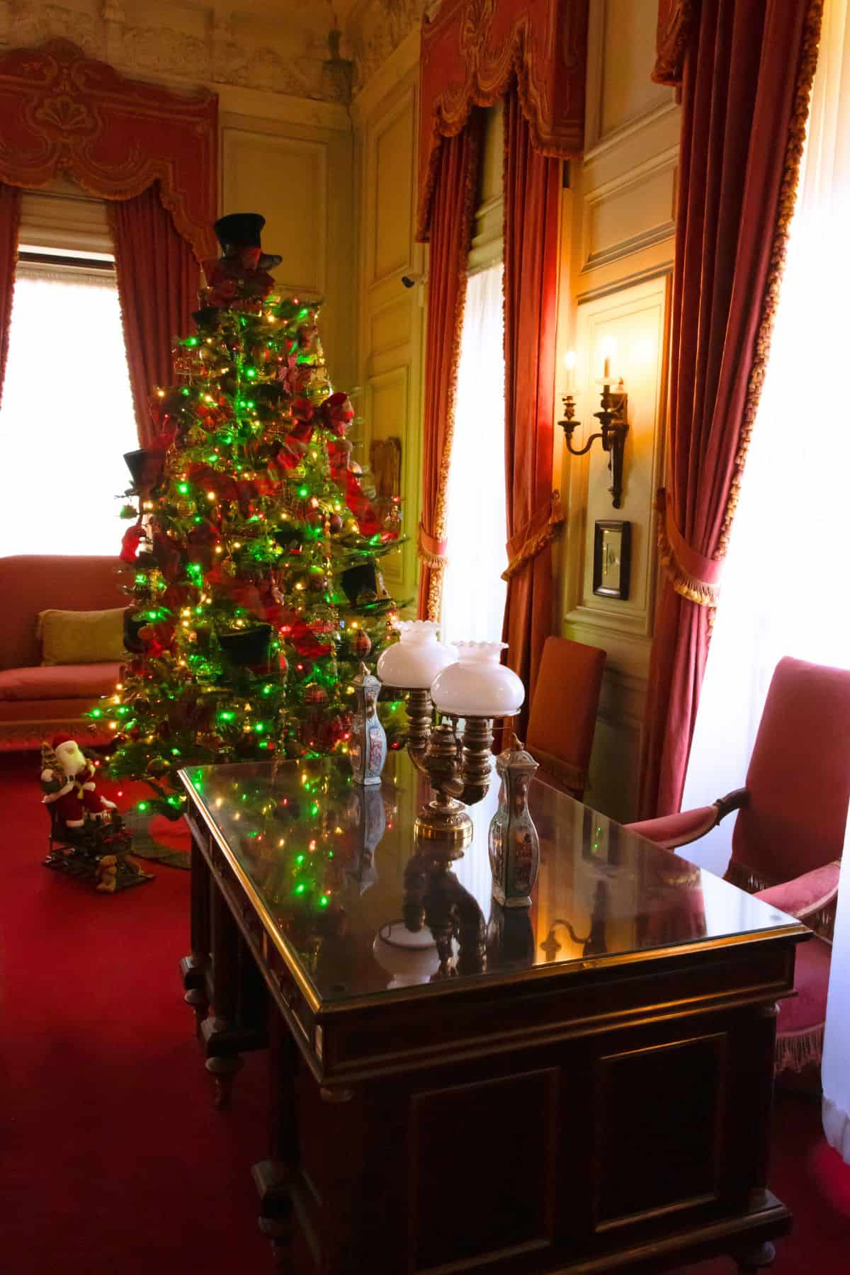 A decorated Christmas tree with green lights stands in a formal room with red curtains, an ornate desk, and antique lamps.
