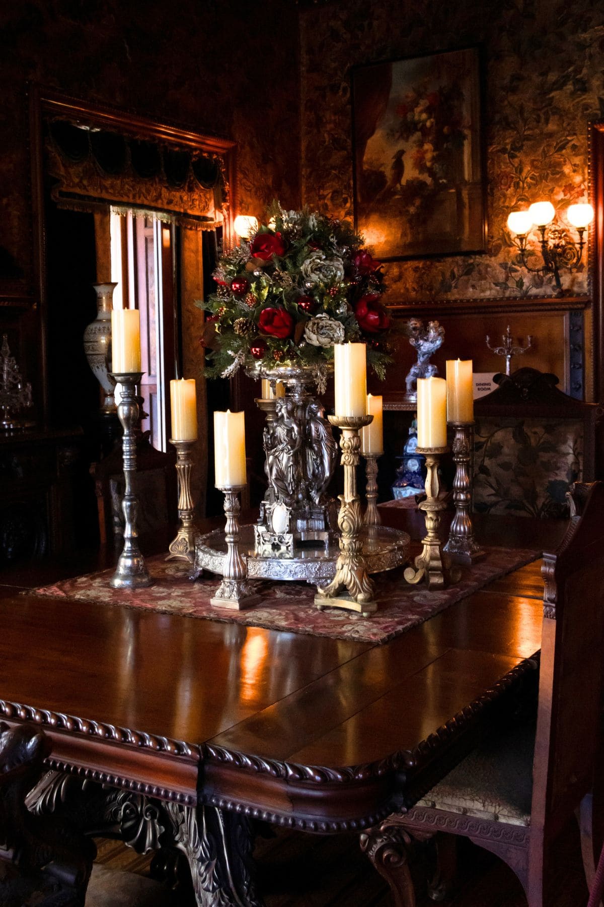 Ornate wooden dining table with gold and silver candle holders, unlit candles, and a floral centerpiece in a dimly lit, vintage-style room.