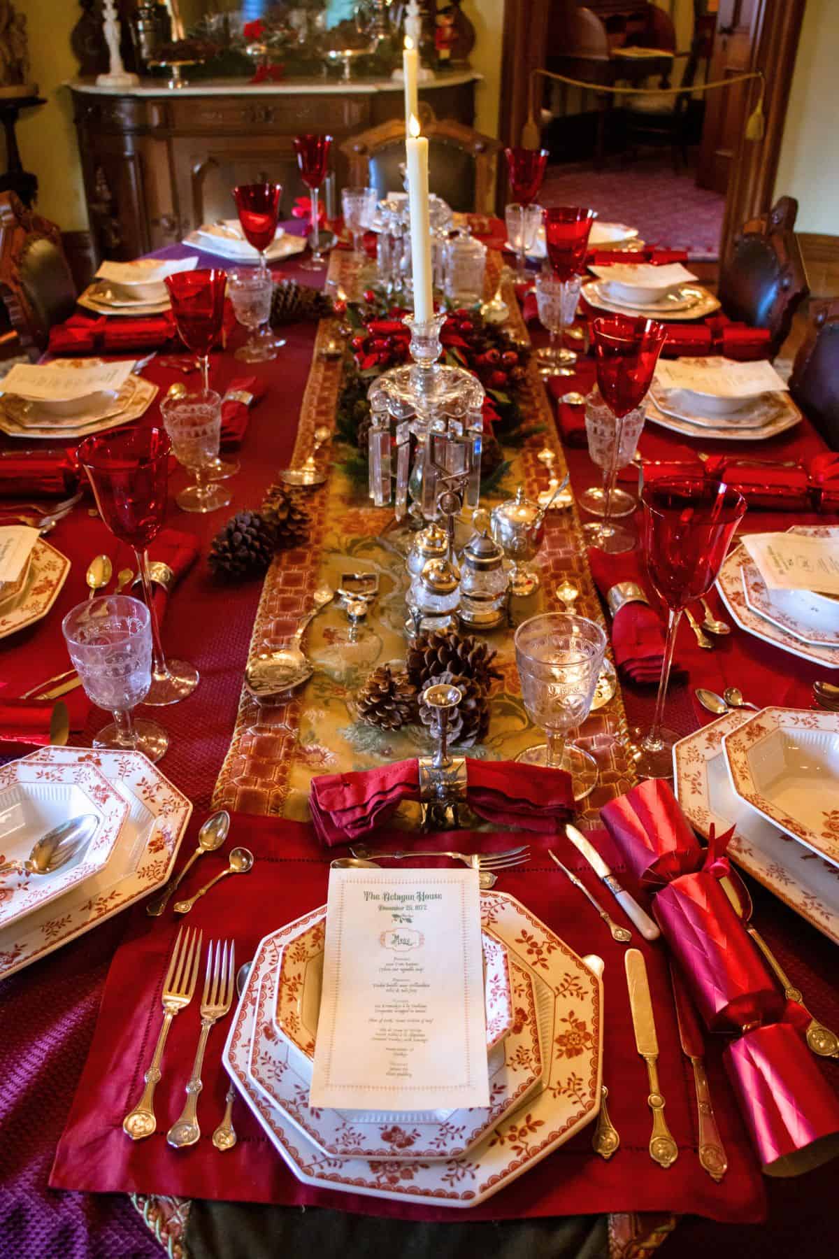A formal dining table set for a holiday meal with red and gold decorations, crystal glasses, silverware, menus, and candles. Pinecones and festive accents run down the center.