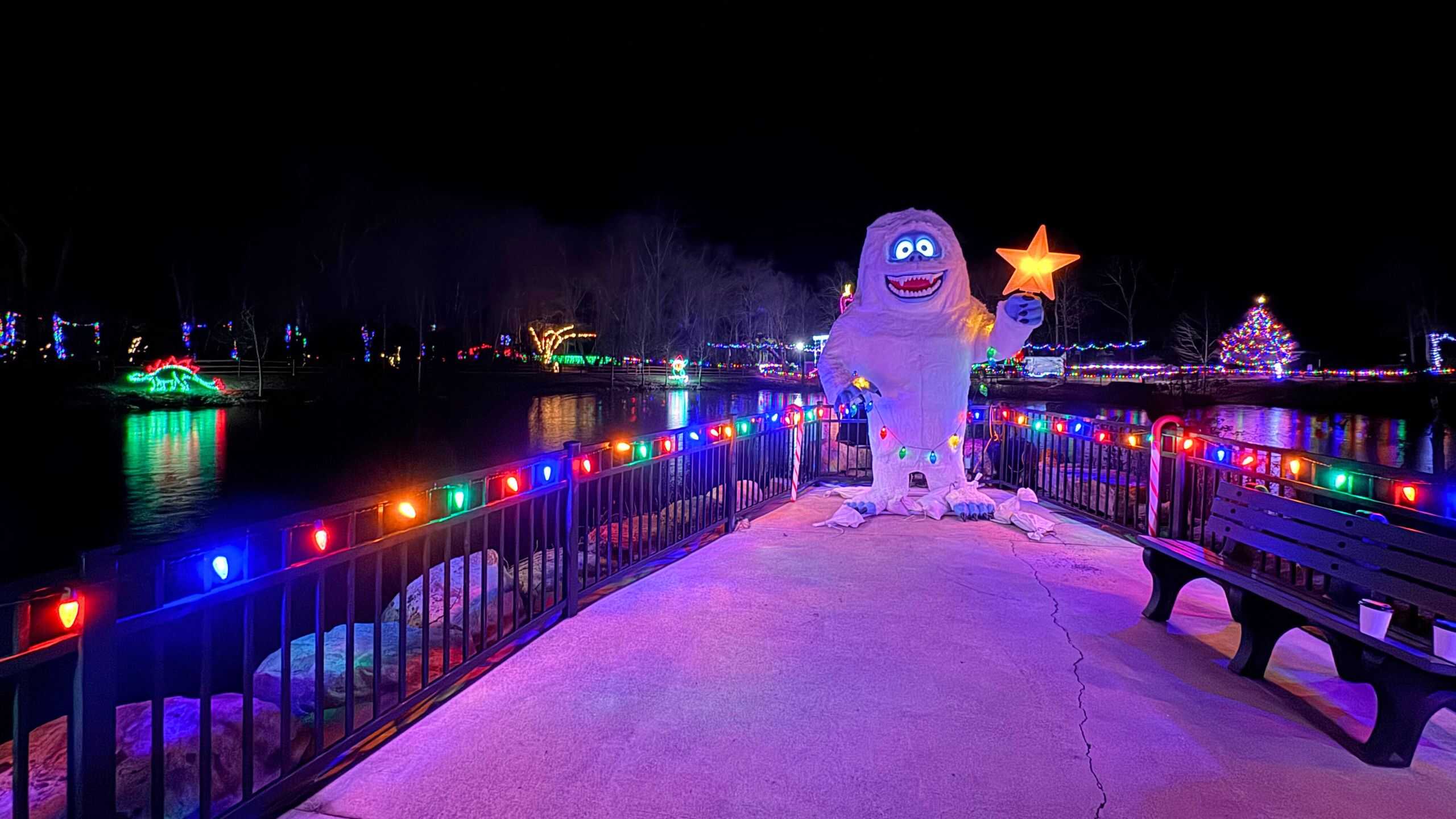 A large abominable snowman figure holds a star on a decorated bridge with colorful holiday lights at night, overlooking a lit-up lake and trees—one of the festive things to do in Connecticut this holiday season.