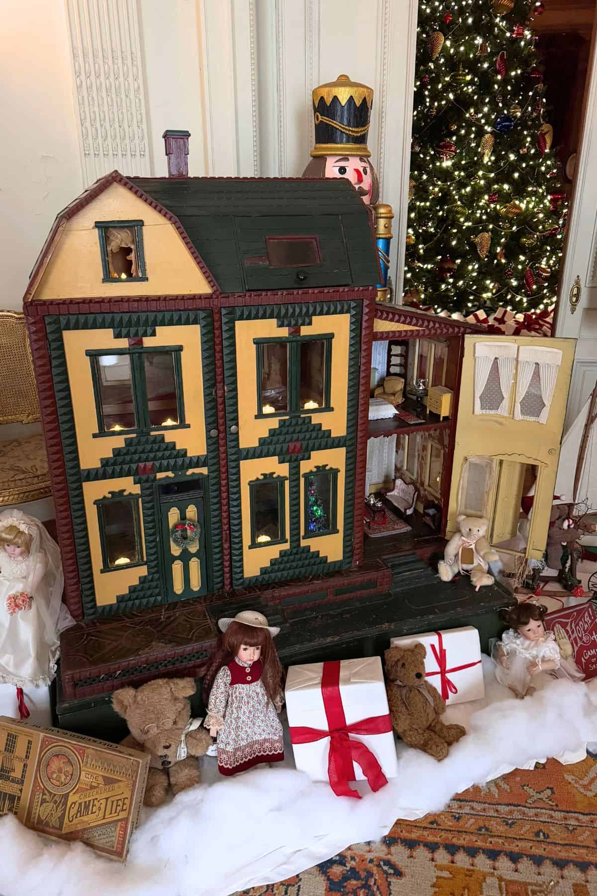 A large vintage dollhouse is displayed with teddy bears, dolls, wrapped gifts, and a decorated Christmas tree in the background.