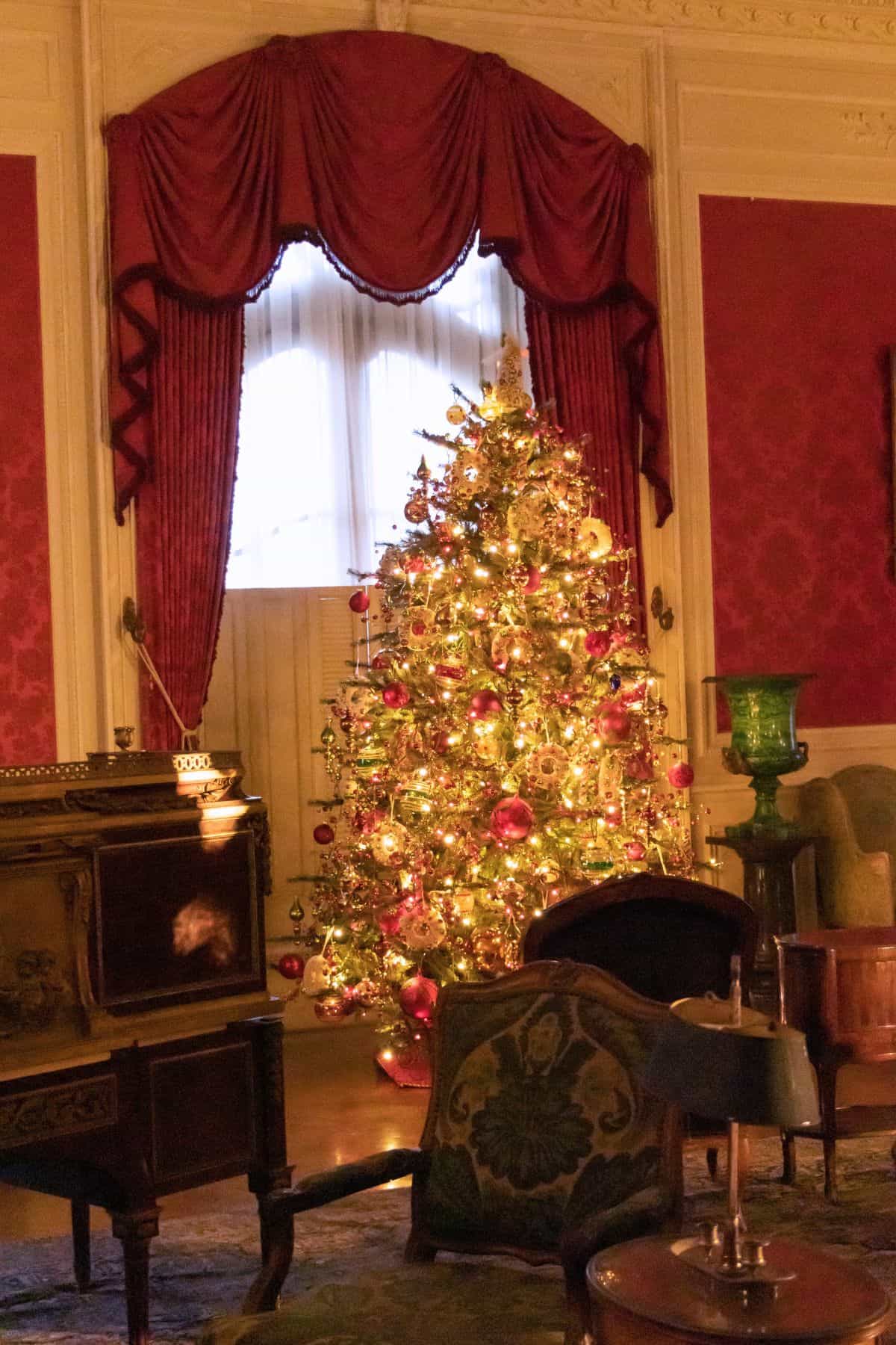A decorated Christmas tree with lights stands in front of a tall window with red drapes in an ornate, vintage-style living room.