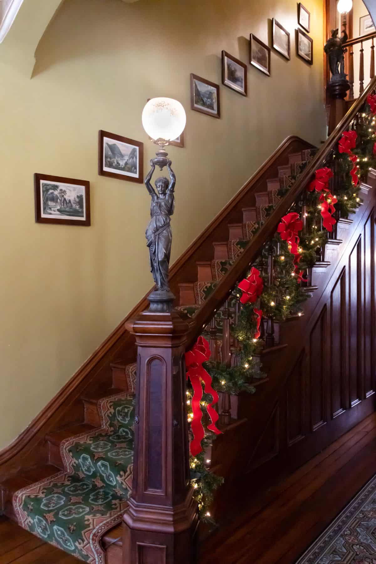 A wooden staircase decorated with garland, red bows, and string lights; framed photos hang on the wall and a statue lamp stands at the base of the stairs.