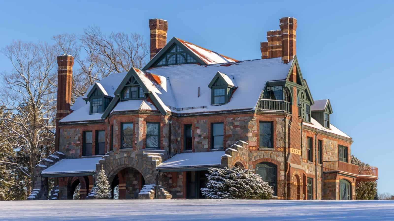 Large, historic brick mansion covered in snow, with multiple chimneys, steep gabled roofs, and arched stone entryway—reminiscent of the grand historic house museums in the northeast—surrounded by snow-covered ground and trees.