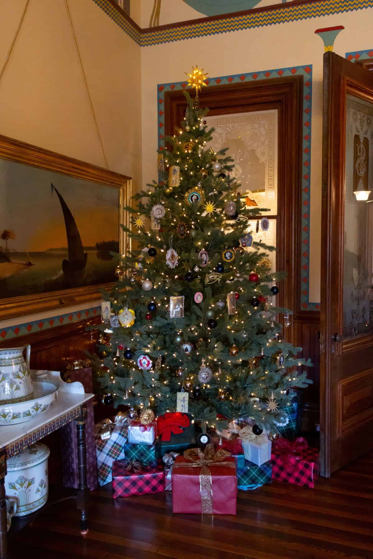 Christmas tree decorated with ornaments and lights, surrounded by wrapped presents, standing in a traditionally furnished room.