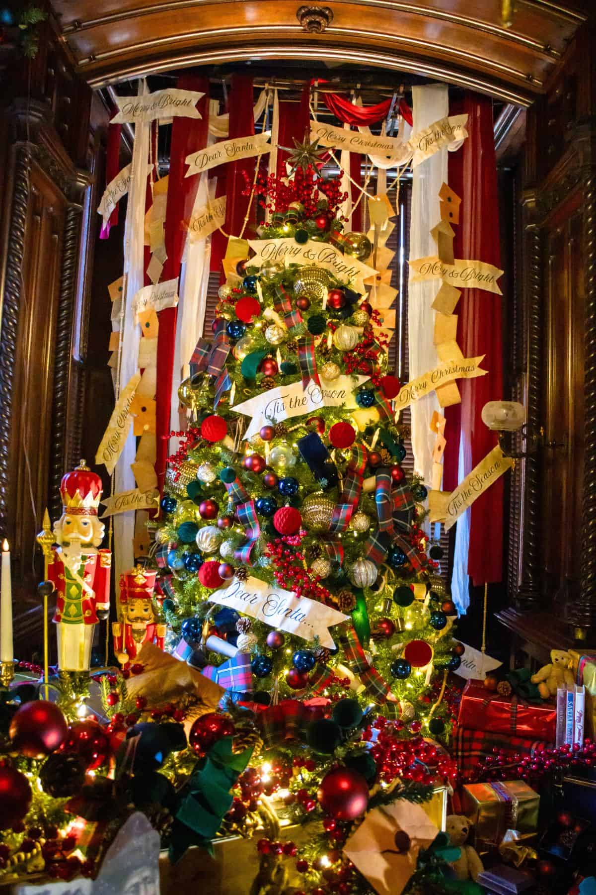 A decorated Christmas tree with red, green, and gold ornaments, surrounded by wrapped gifts, with festive banners and nutcracker figures in a wooden-paneled room.