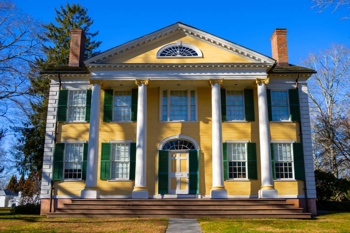 A large yellow colonial-style house with white columns, green shutters, and a prominent central doorway, set against a clear blue sky.