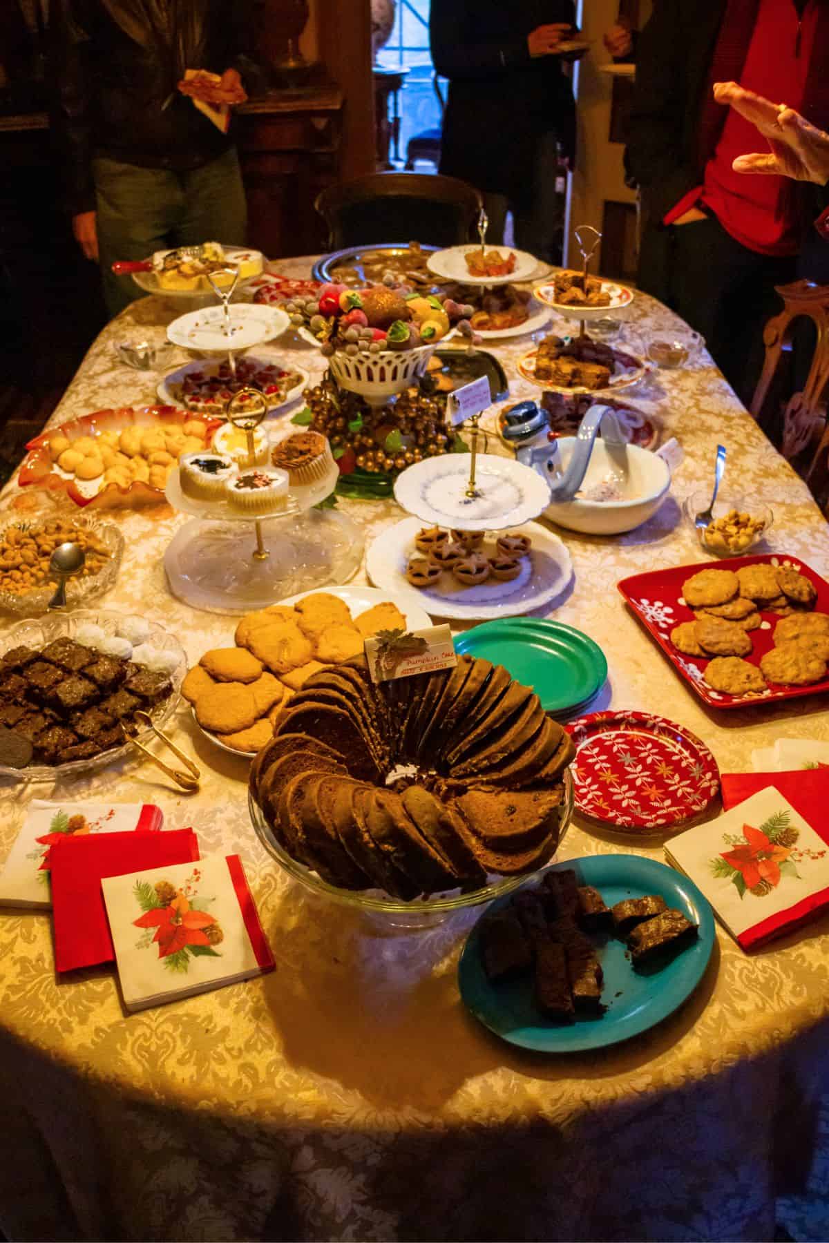 A table covered with a variety of cookies, brownies, and desserts on plates and trays, surrounded by people standing.