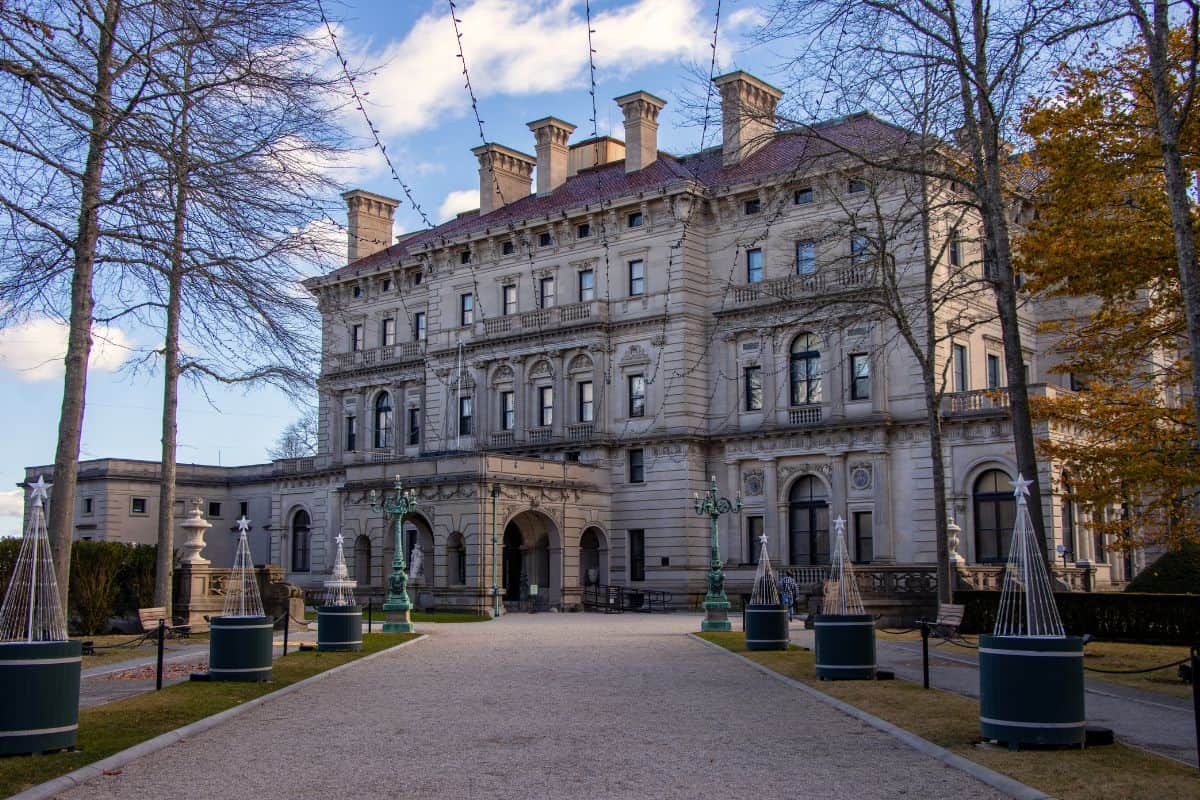 A large, ornate stone mansion with arched entryways and decorative columns, viewed from a pathway lined with trees and light fixtures.