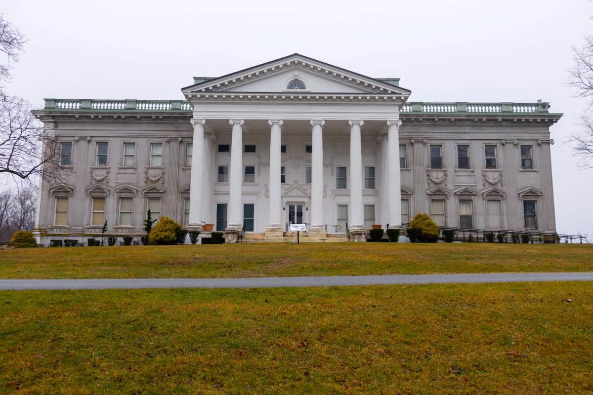 Large, white, neoclassical mansion with tall columns, symmetrical windows, and a triangular pediment, set on a grassy lawn under an overcast sky.