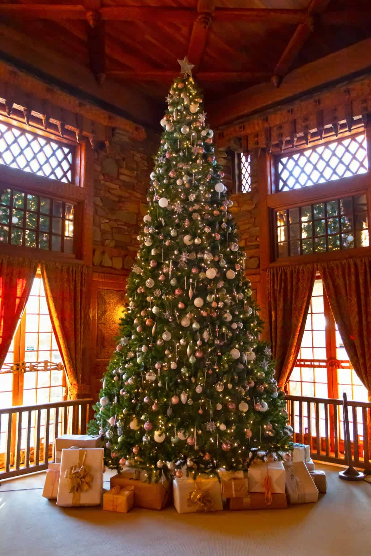 A large decorated Christmas tree with white ornaments and lights stands in front of windows, surrounded by wrapped presents in a wooden room.