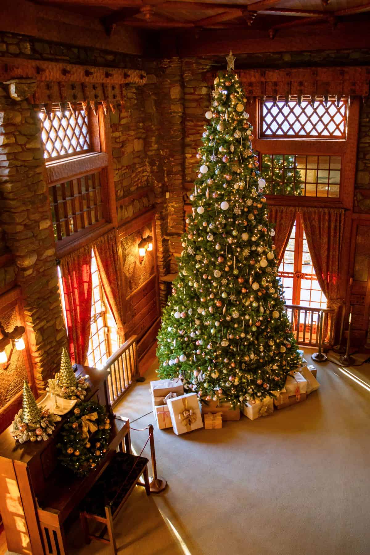 A tall, decorated Christmas tree stands in a warmly lit room with stone walls, wooden accents, and wrapped gifts arranged at its base.