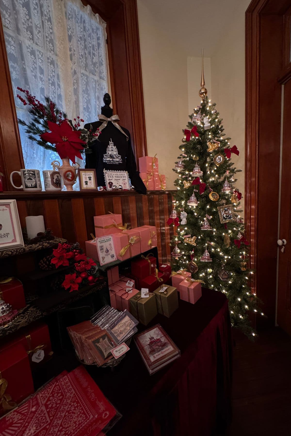 A decorated Christmas tree with white lights and pink ornaments stands near a table displaying wrapped presents, cards, and festive decorations in a warmly lit room.