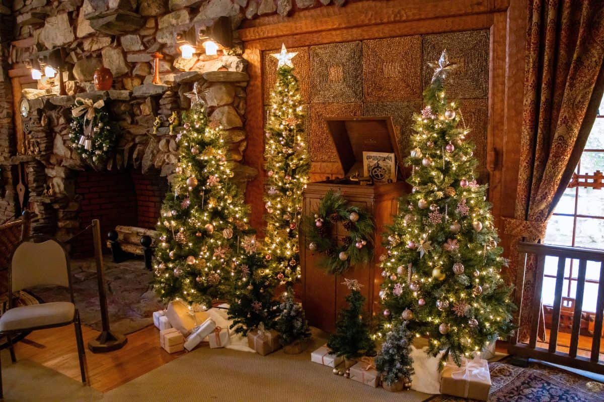 Four decorated Christmas trees with white lights and ornaments stand near wrapped gifts in a cozy room with a stone fireplace and wood-paneled walls.