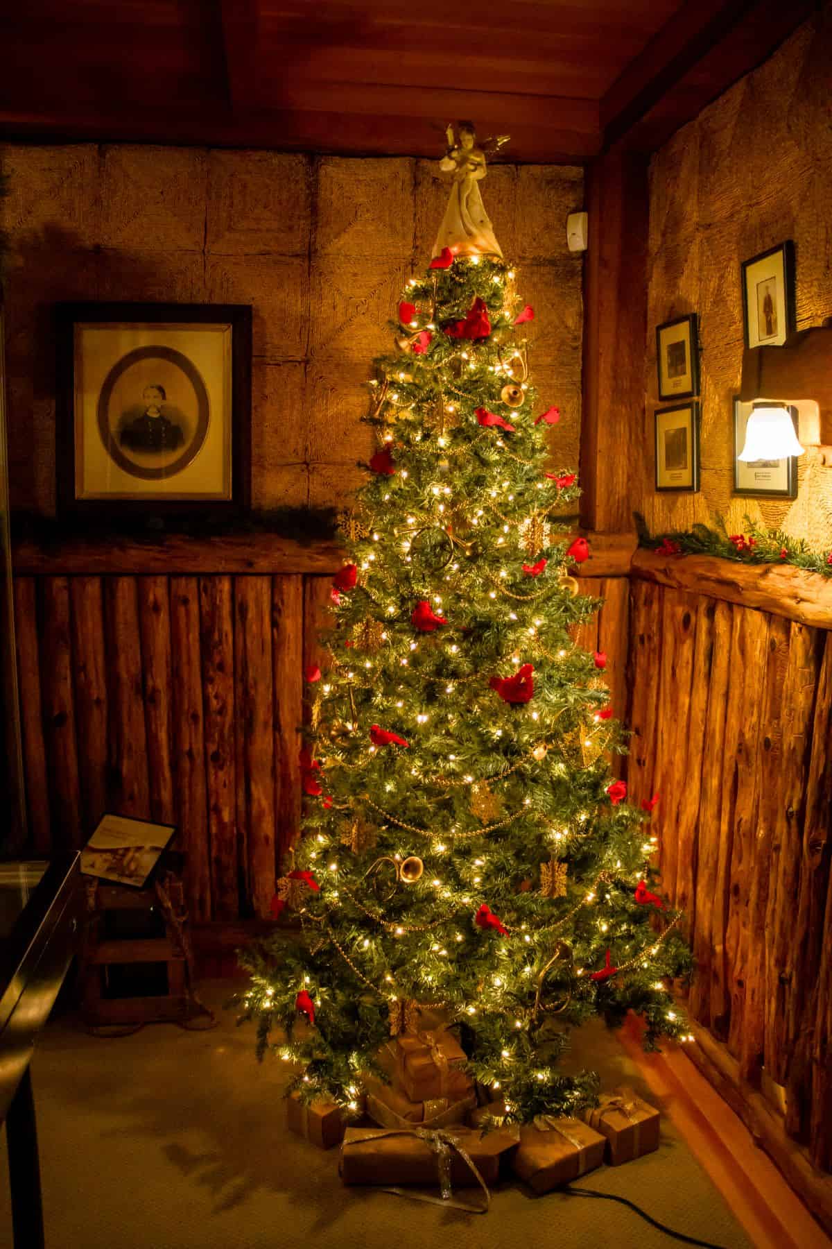 A decorated Christmas tree with lights and red ornaments stands in a rustic wooden room, surrounded by wrapped gifts at its base.