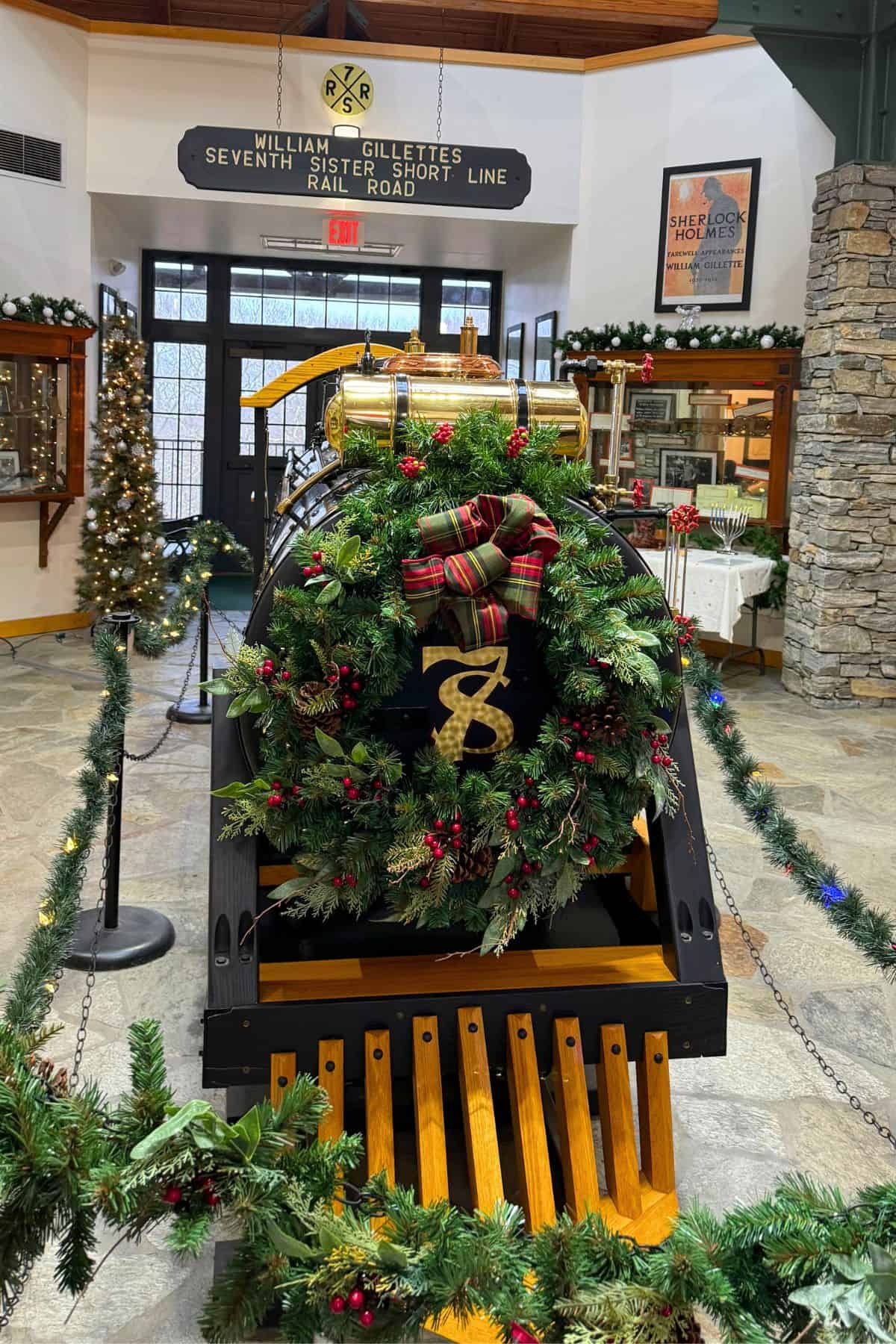 A vintage locomotive decorated with a festive wreath and ribbon is displayed indoors at the William Gillette's Seventh Sister Short Line Railroad exhibit.