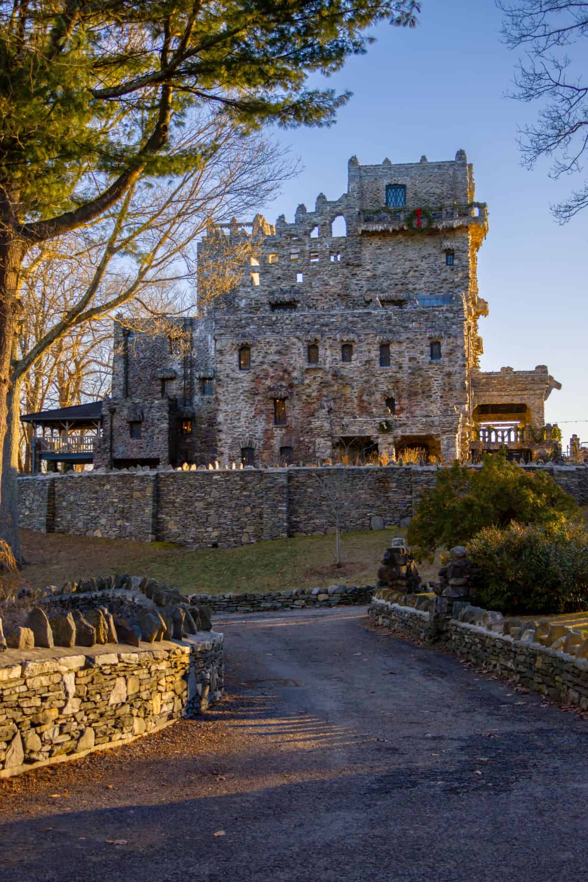 A stone castle with a rugged, uneven facade stands behind a stone wall, surrounded by trees and a winding path in the foreground.