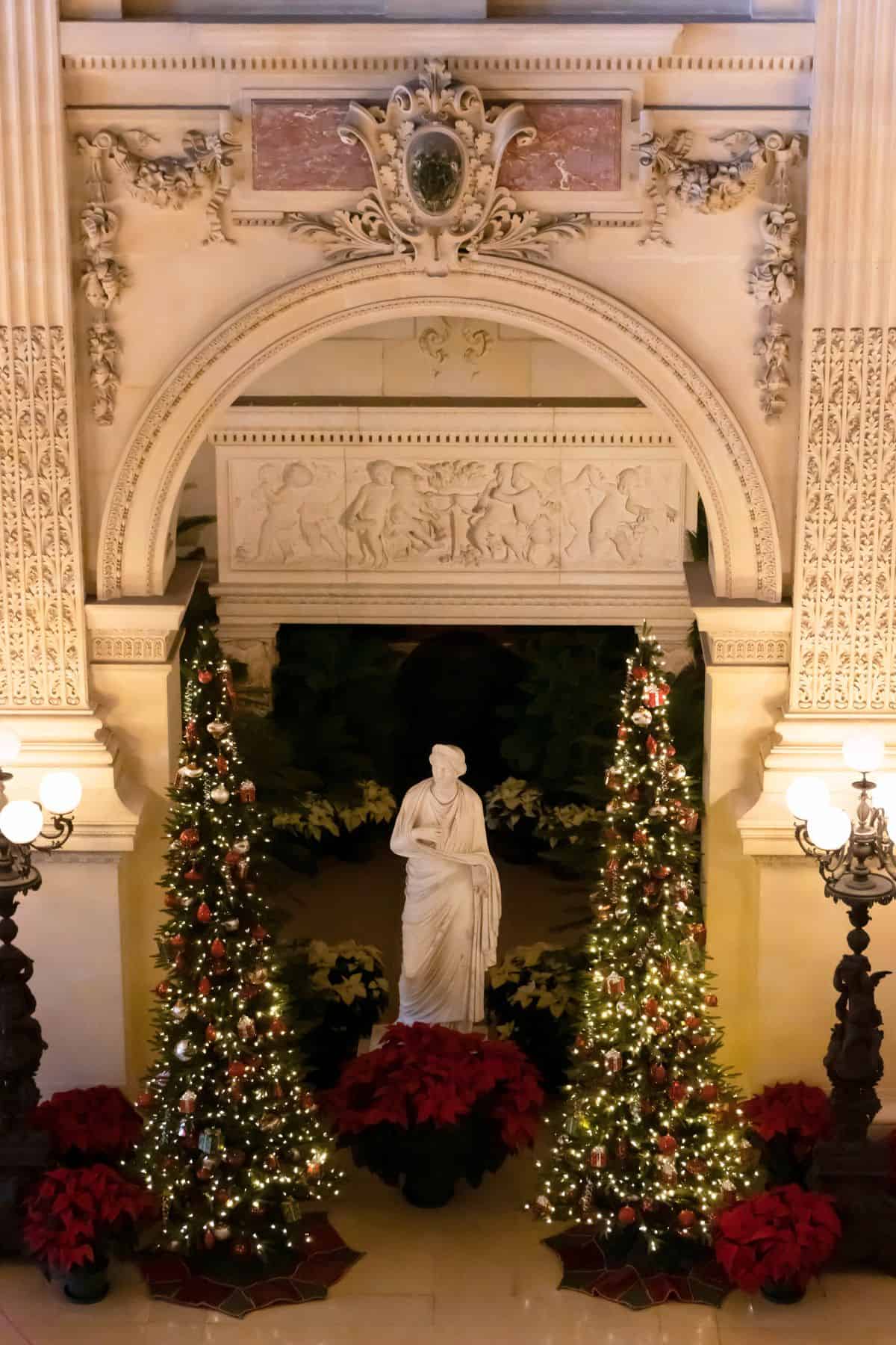 A marble statue stands between two decorated Christmas trees under an ornate archway, with red poinsettias and architectural details in the background.