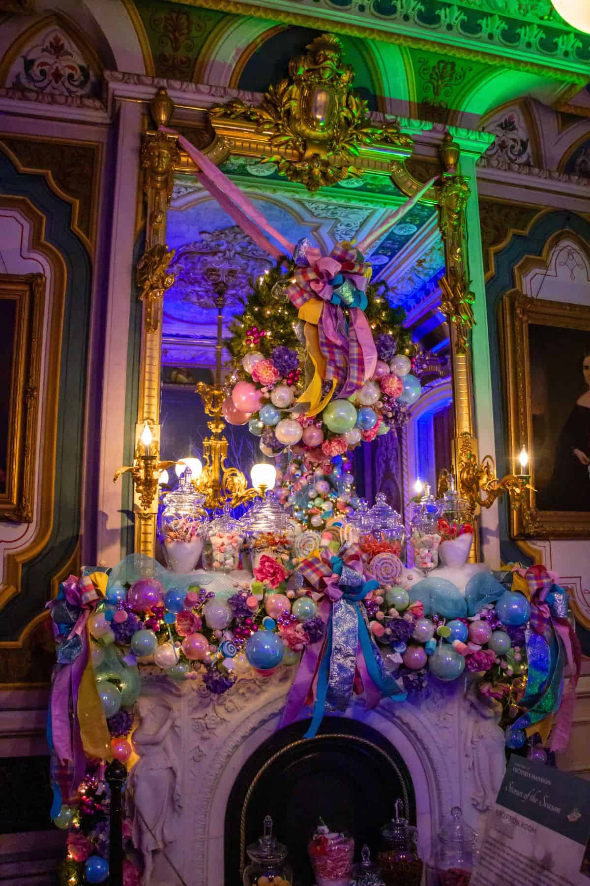 A lavishly decorated fireplace with colorful ornaments, pastel ribbons, and a festive wreath, set in an ornate room with gold-framed mirrors and portraits.