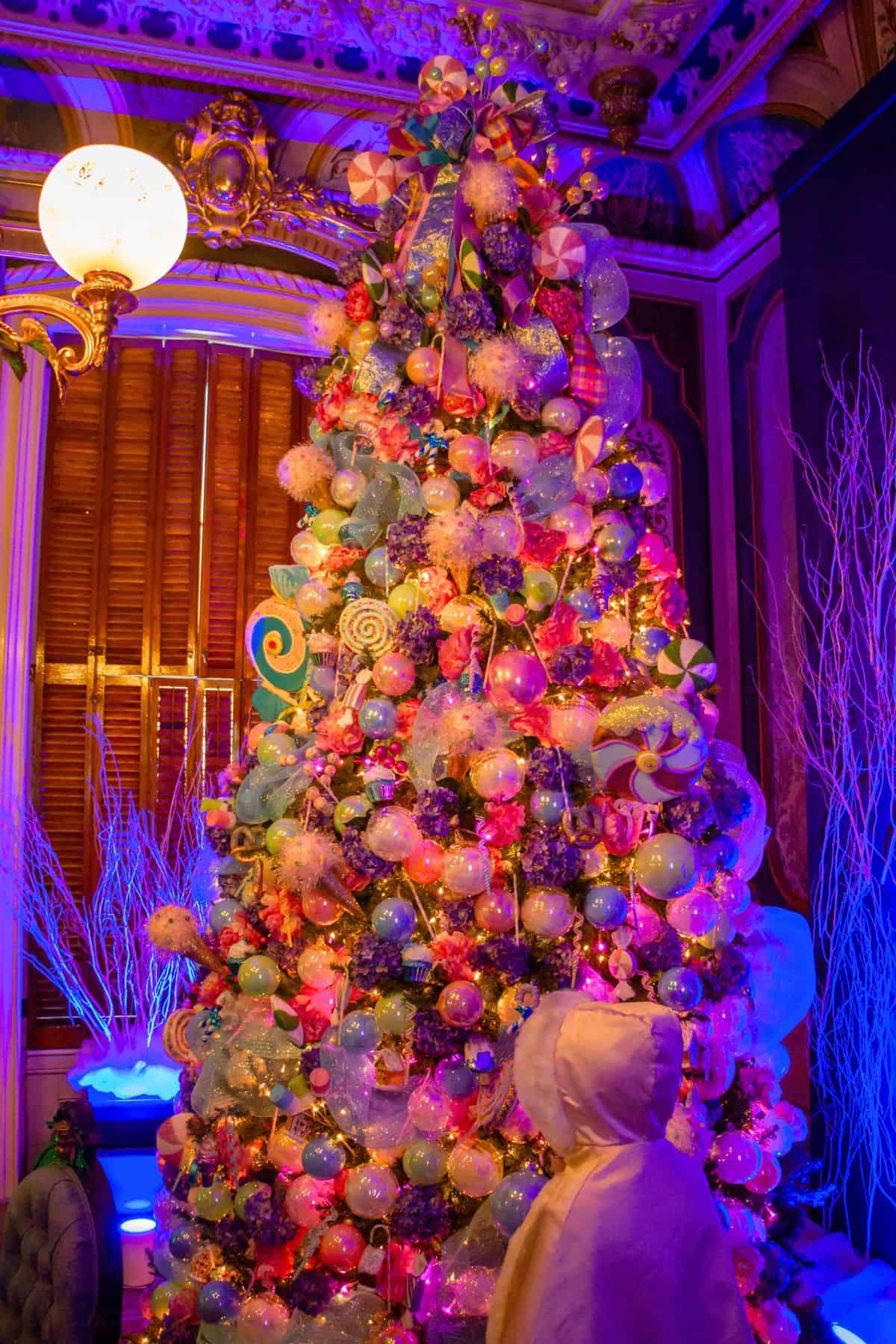 A person in a white hooded coat stands in front of a brightly lit Christmas tree decorated with colorful ornaments and candy-themed decorations in an ornate room.