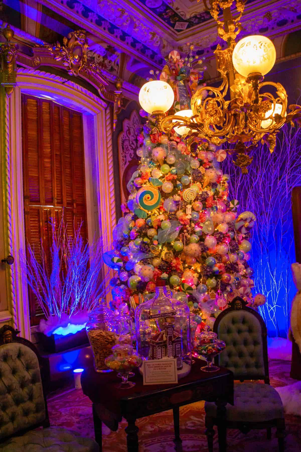 A brightly lit Christmas tree decorated with colorful ornaments stands in an ornate room with vintage furniture, under a decorative chandelier.