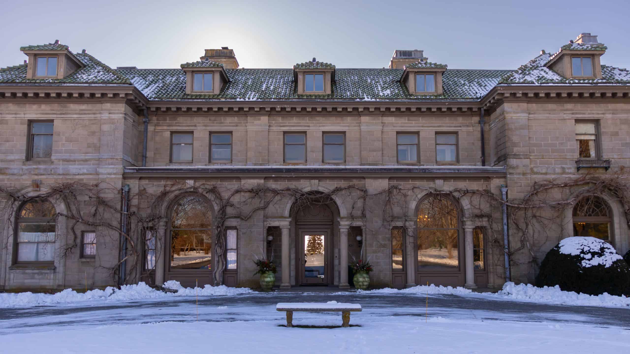 Large stone mansion with arched windows and doors, snow on the roof and ground, barren vines on the facade, and a bench in front—perfect for enjoying festive things to do in Connecticut this holiday season as sunlight shines behind the building.