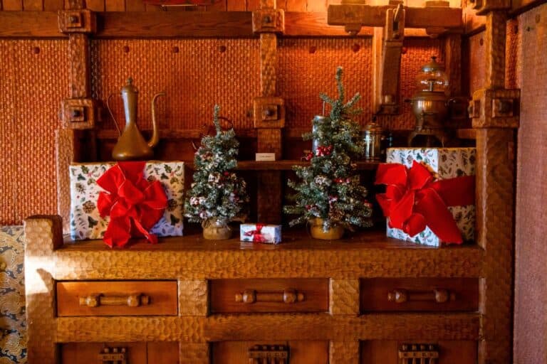 A wooden cabinet displays two small decorated Christmas trees, wrapped gifts with red bows, and brass pitchers against a woven-patterned wall.
