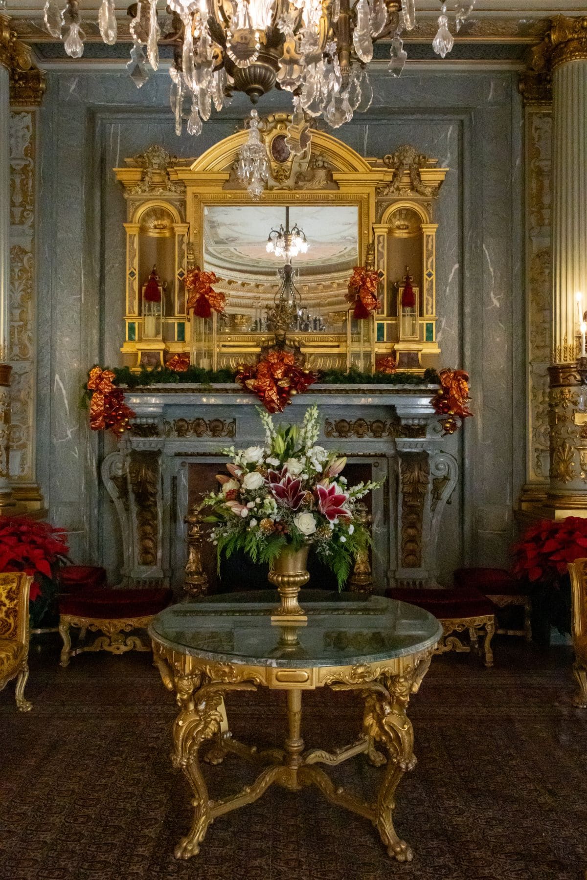 Ornate room with a marble fireplace decorated with red and gold garlands, a floral arrangement on a round table, and a chandelier overhead.