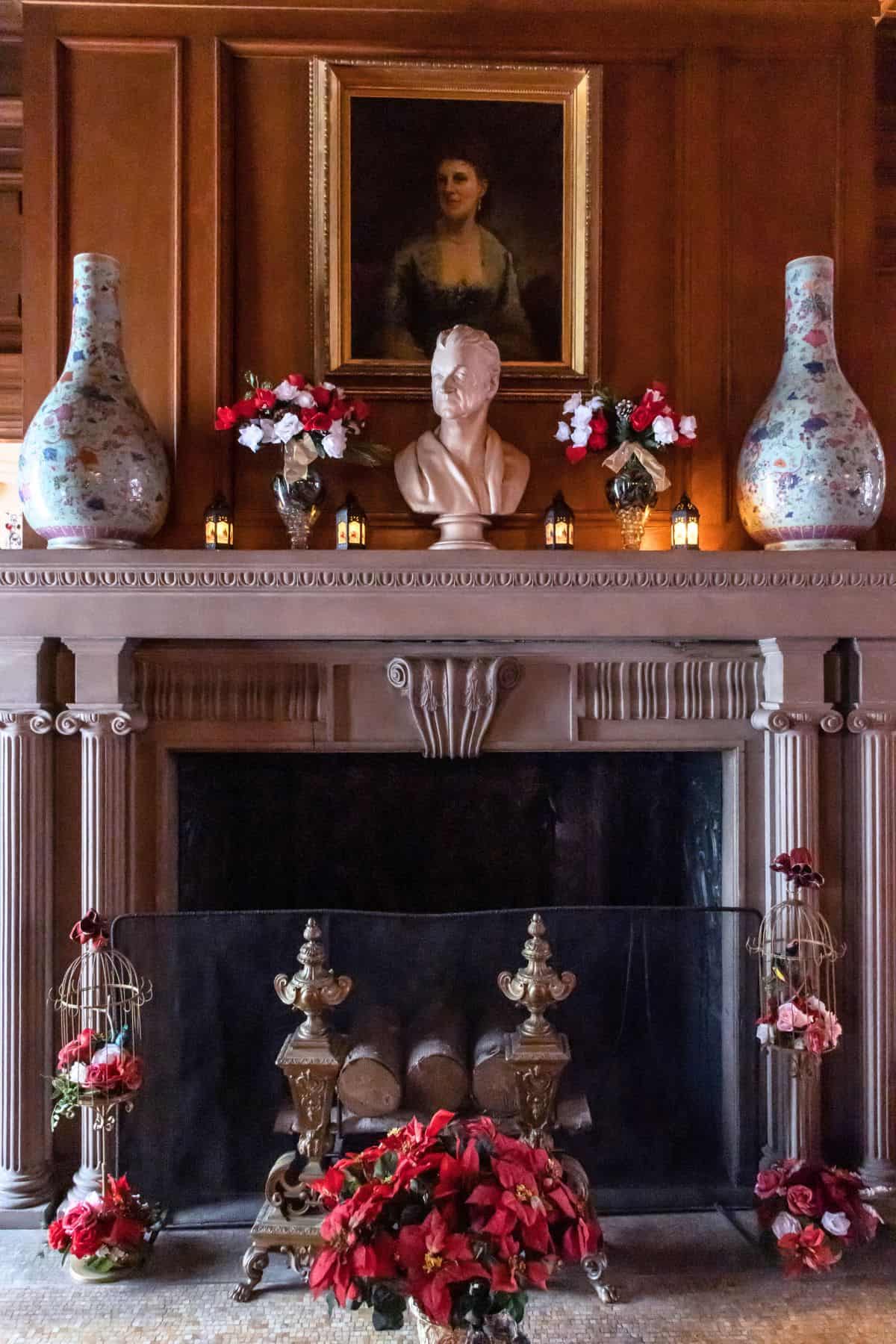 A decorated fireplace features a bust, floral arrangements, two large vases, and a portrait of a woman above the mantel.
