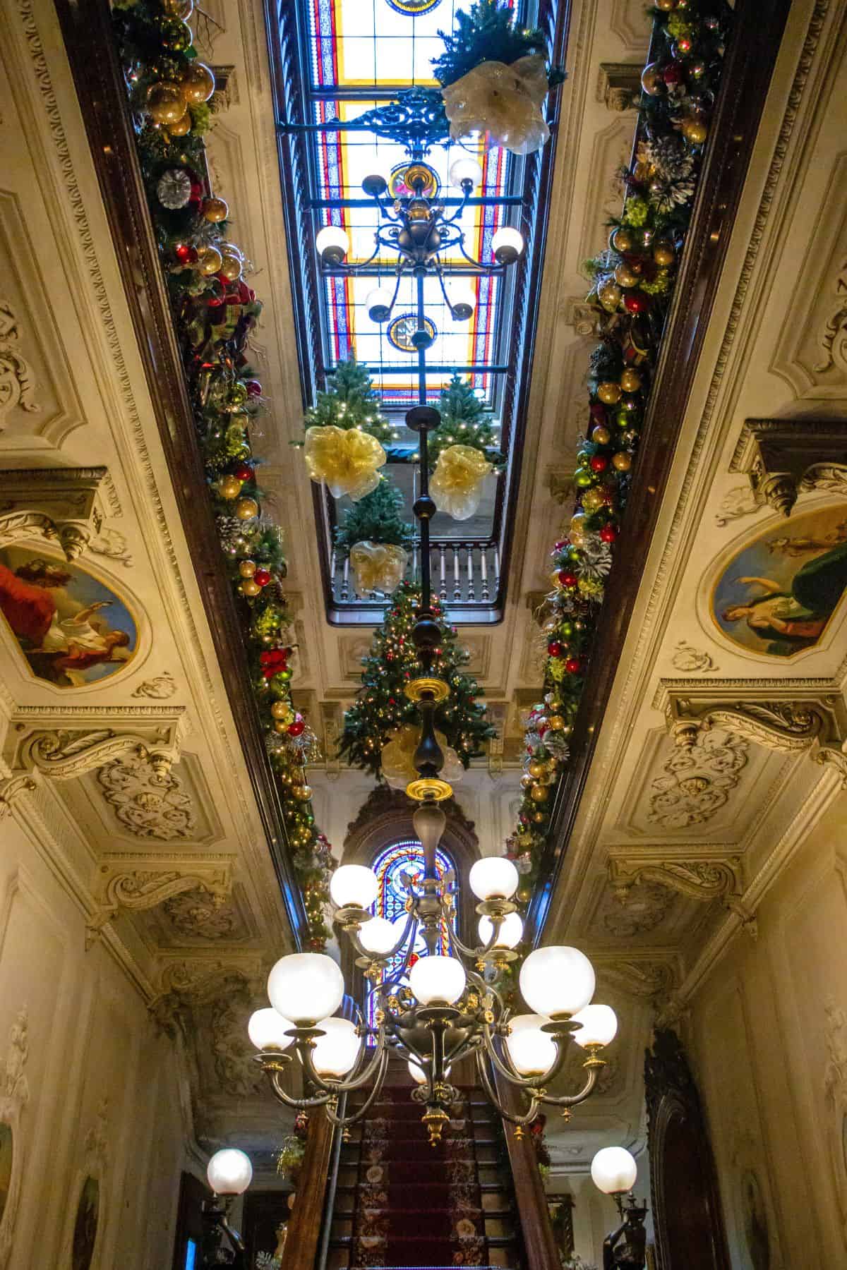 A grand staircase with ornate railings, a red carpet, and a large chandelier, decorated with garlands and Christmas ornaments under a stained glass ceiling.