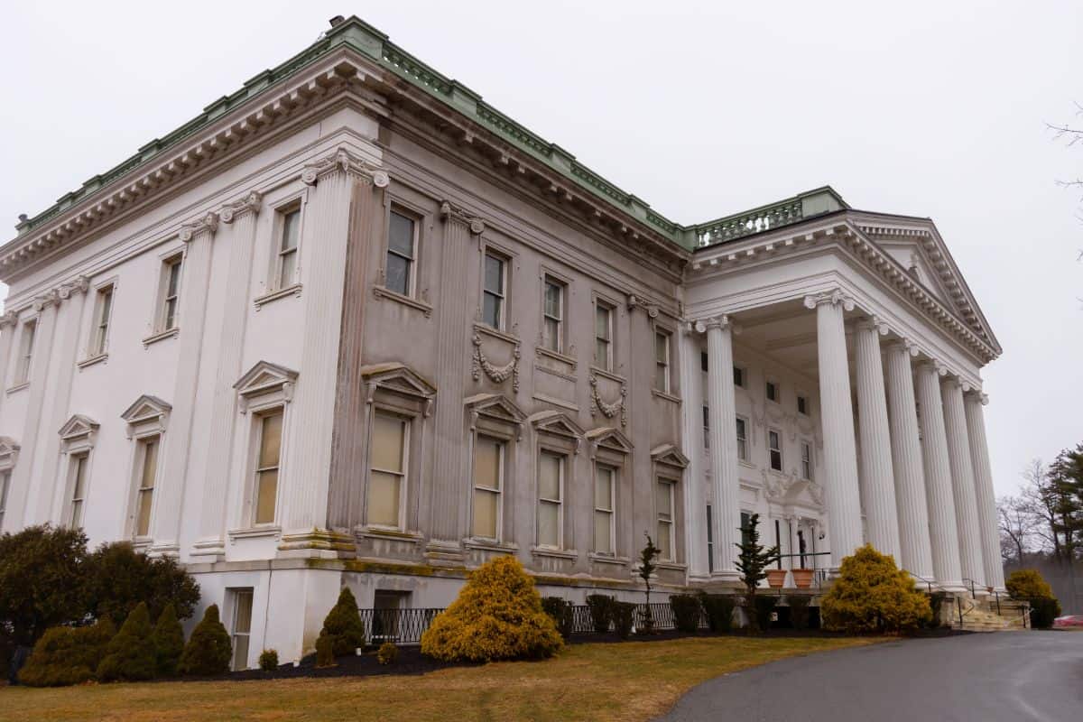 Large, white, neoclassical building with tall columns, multiple windows, and weathered exterior; overcast sky and trimmed bushes in the foreground.