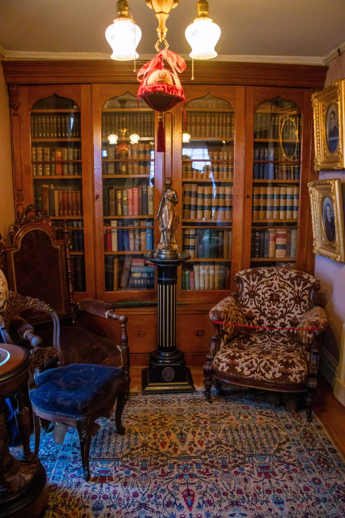 A vintage library room with a large wooden bookshelf filled with books, ornate chairs, a patterned rug, framed portraits, and a decorative lamp and statue.