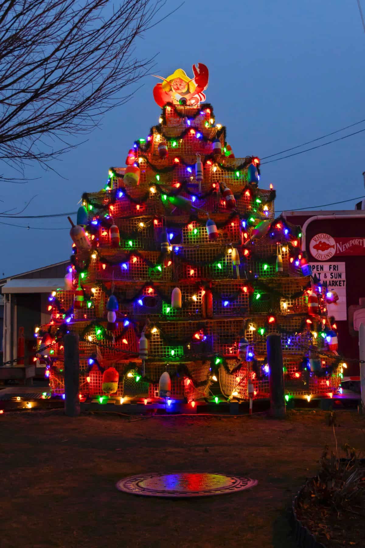 A large outdoor Christmas tree made of stacked lobster traps decorated with multicolored lights, buoys, and a Santa Claus figure on top, displayed at dusk.